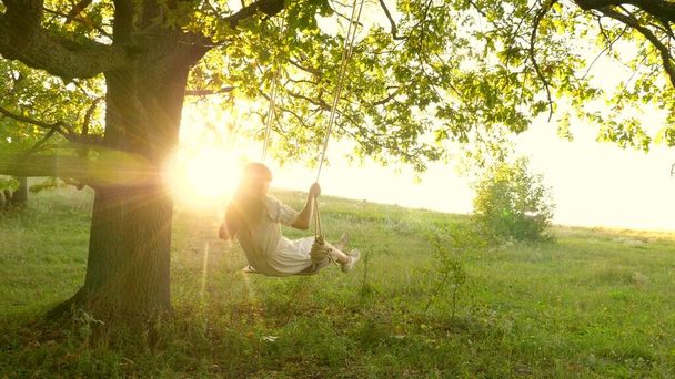 Ένα νεαρό κορίτσι αιωρείται σε μια κούνια σε ένα κλαδί βελανιδιάς στον ήλιο. Όνειρα πτήσης. Χαρούμενη παιδική ιδέα. Όμορφο κορίτσι με λευκό φόρεμα στο πάρκο. έφηβος κορίτσι απολαμβάνει πτήση σε ταλάντευση το βράδυ του καλοκαιριού στο δάσος - Φωτογραφία, εικόνα