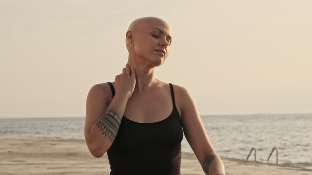 ruhige attraktive Sportlerin mit Glatze, die ihren Hals mit geschlossenen Augen berührt und danach wegsieht, während sie im Freien am Meer sitzt - Filmmaterial, Video