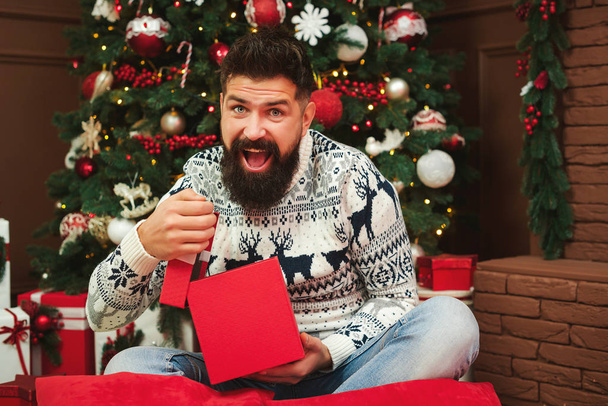 Um hipster surpreso abre um presente de Natal. Homem barbudo feliz sentado perto da árvore de Natal. Homem desfrutando feriados de Natal em casa
 - Foto, Imagem