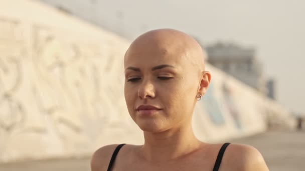 Close up view of pleased attractive bald sports woman sitting on mat with closed eyes near the sea outdoors - Filmagem, Vídeo