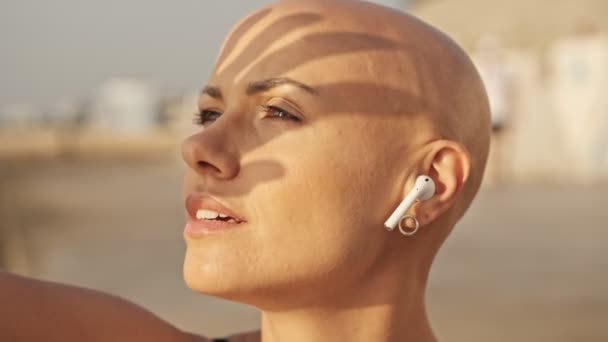 Close up view of Pleased attractive bald sports woman in earphones with finger shadow on the face listening music and looking away while sitting near the sea outdoors - Filmagem, Vídeo