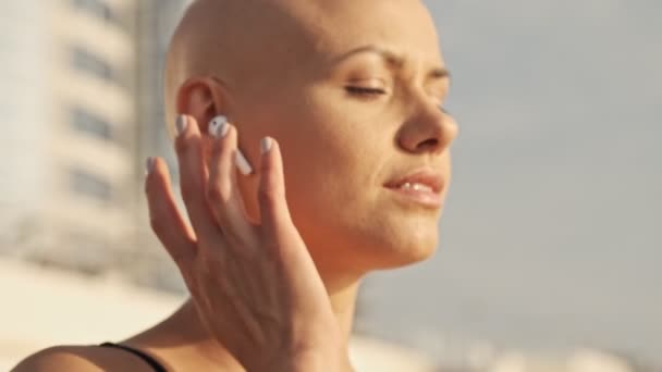 Side view of Pleased attractive bald sports woman touching earphone and becoming happy while sitting near the sea outdoors - Záběry, video