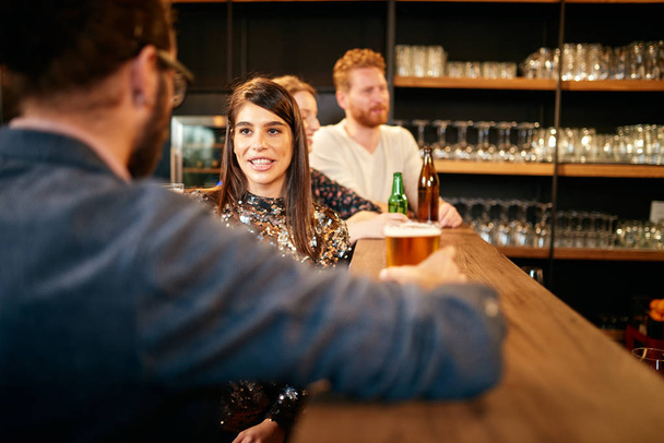 Вид сзади на красивого кавказца, опирающегося на барную стойку, пьющего пиво и флиртующего с женщиной, с которой только что познакомился. Интерьер паба
. - Фото, изображение