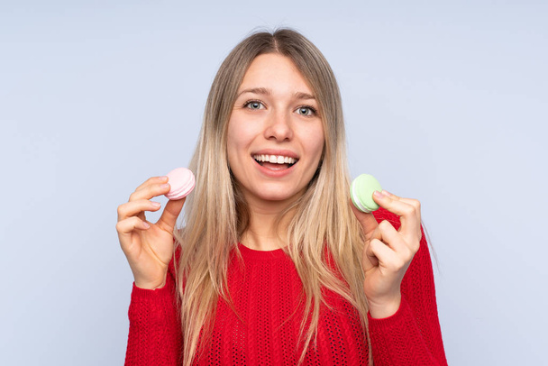Young blonde woman over isolated blue background holding colorful French macarons - Fotografie, Obrázek