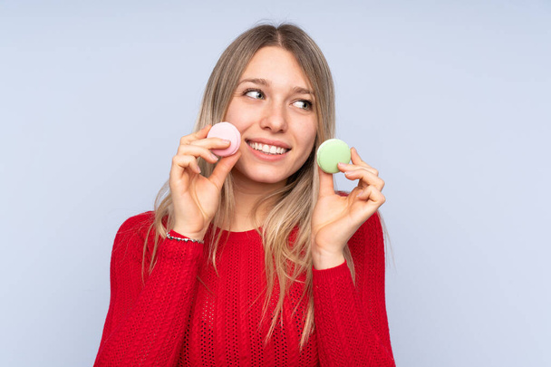 Young blonde woman over isolated blue background holding colorful French macarons and looking up - Foto, afbeelding