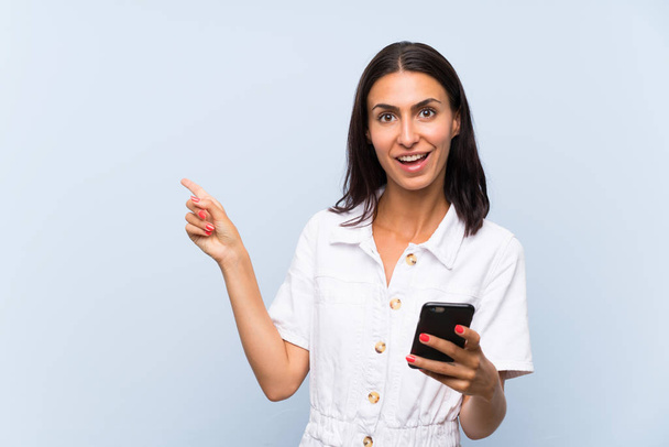 Young woman with a mobile phone over isolated blue wall surprised and pointing finger to the side - Photo, image