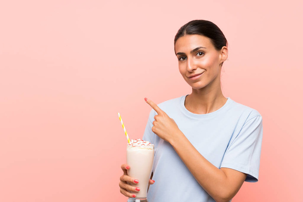 Soyutlanmış pembe arka planda çilekli milkshake 'i olan genç bir kadın bir ürünü sunmak için kenarı işaret ediyor. - Fotoğraf, Görsel