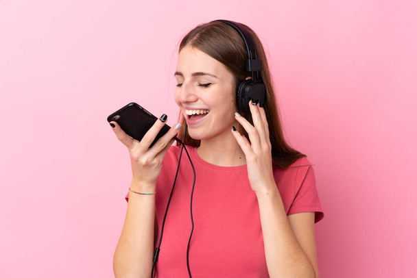Soyutlanmış pembe arka planı olan genç bir kadın cep telefonuyla müzik dinliyor ve şarkı söylüyor. - Fotoğraf, Görsel