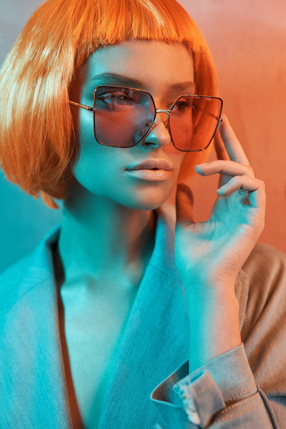 portrait of a beautiful girl in orange glasses on neon background with nude makeup and short orange hair she dressed in jeans jacket and looks in the right - Foto, immagini