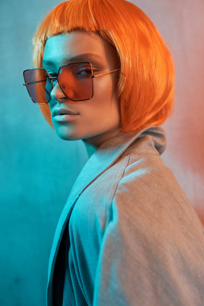 портрет красивої дівчини в помаранчевих окулярах на неоновому тлі з оголеним макіяжем і коротким помаранчевим волоссям, яке вона одягнула в джинсову куртку і дивиться в камеру
 - Фото, зображення
