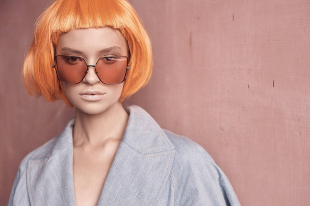 portrait of a beautiful girl in orange glasses on a pink background with nude makeup and short orange hair she dressed in jeans jacket and looks in the camera - Foto, afbeelding