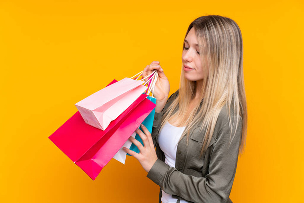 Young blonde woman over isolated yellow background holding shopping bags - Photo, Image