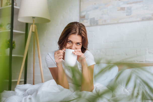 selective focus of young woman looking at camera while holding cup with tea in bed  - Foto, Imagem