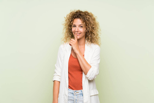 Young blonde woman with curly hair over isolated green background doing silence gesture - Photo, Image