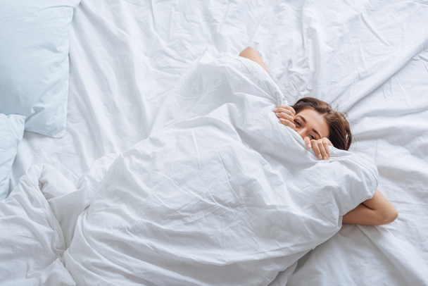 Draufsicht der jungen Frau, die Gesicht mit Decke bedeckt, während sie sich im Bett ausruht  - Foto, Bild