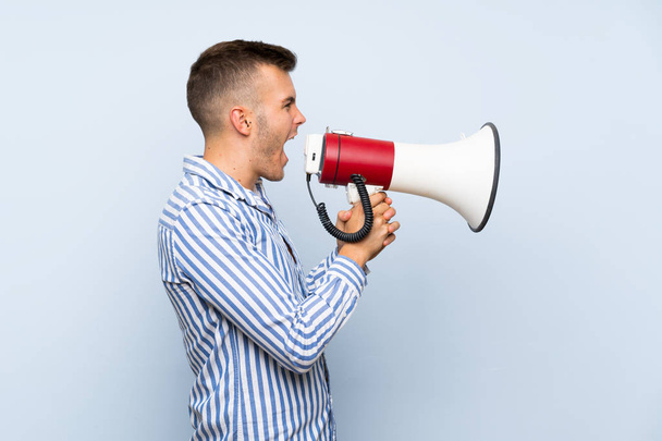 Young handsome blonde man over isolated blue background shouting through a megaphone - 写真・画像