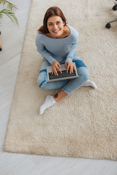 top view of cheerful blogger using laptop while sitting on carpet  - Фото, зображення