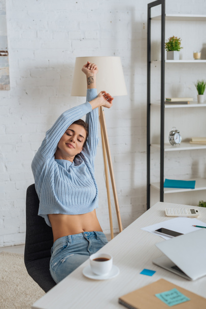 attractive young woman dreaming while stretching near gadgets on table  - Photo, Image