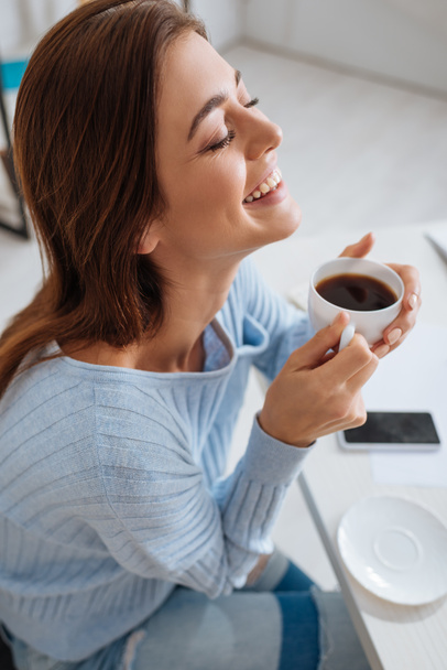 veselá mladá žena se usmívá, zatímco drží šálek kávy  - Fotografie, Obrázek