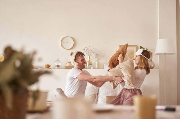 Ζευγάρι ερωτευμένο να γελάει και να παίζει στο κρεβάτι. Άντρας και γυναίκα πολεμούν μαξιλάρια. Νεαρό ευτυχισμένο ζευγάρι κτυπά τα μαξιλάρια στο κρεβάτι σε ένα υπνοδωμάτιο στο σπίτι - Φωτογραφία, εικόνα