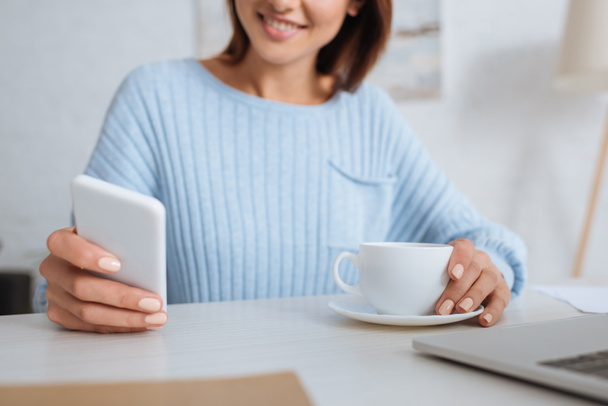 Ausgeschnittene Ansicht einer fröhlichen Frau mit Smartphone in der Nähe von Tasse mit Kaffee  - Foto, Bild