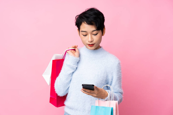 Jovencita asiática sobre fondo rosa aislado sosteniendo bolsas de compras y escribiendo un mensaje con su teléfono celular a un amigo - Foto, Imagen