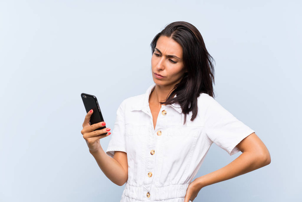 Jonge vrouw met een mobiele telefoon over geïsoleerde blauwe muur - Foto, afbeelding