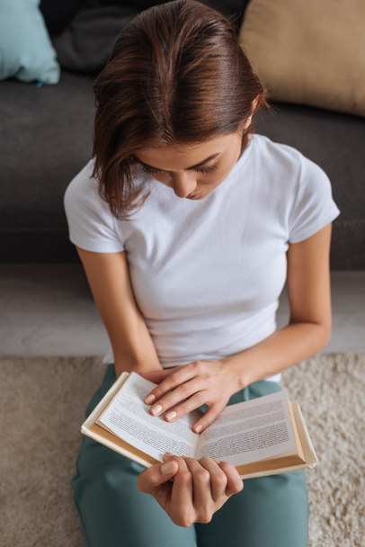 bovenaanzicht van vrouw boek lezen terwijl zitten op tapijt in de buurt van bank  - Foto, afbeelding