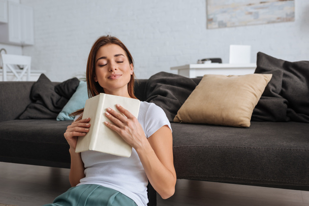 dreamy girl holding book while sitting in living room  - Photo, Image