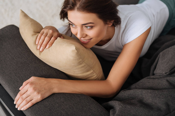 γενικά θέα χαμογελαστή γυναίκα ψύξη, ενώ βρίσκεται στον καναπέ κοντά στο μαξιλάρι  - Φωτογραφία, εικόνα