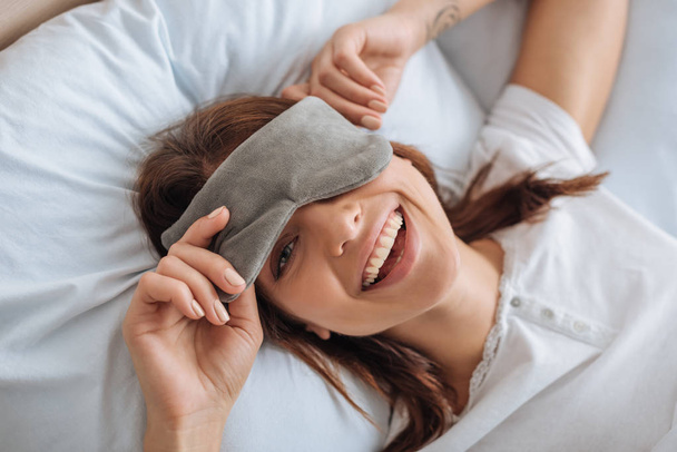 Blick von oben auf eine glückliche junge Frau mit Augenmaske, die im Schlafzimmer ruht  - Foto, Bild