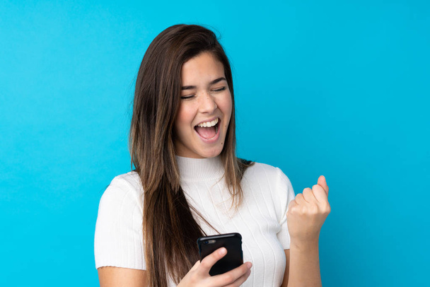Teenager dívka přes izolované modré pozadí s telefonem ve vítězné pozici - Fotografie, Obrázek