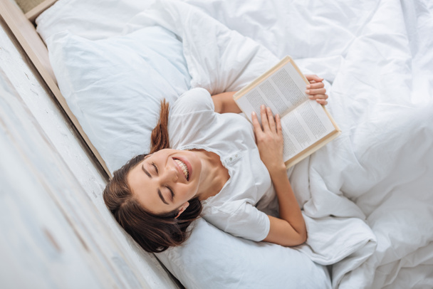 top view of happy girl holding book while chilling in bed  - Photo, Image