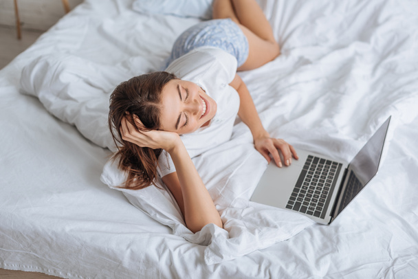 wesoła kobieta korzystająca z laptopa podczas chłodzenia w sypialni  - Zdjęcie, obraz