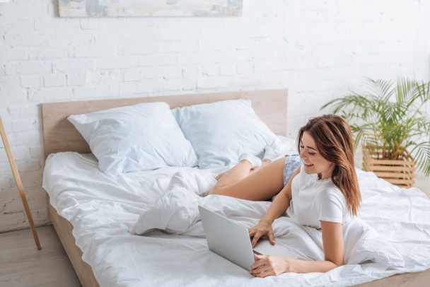 cheerful young woman using laptop while chilling in bedroom  - Fotografie, Obrázek