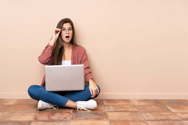 tiener student meisje zitten op de vloer met een laptop met bril en verrast - Foto, afbeelding