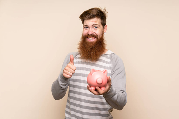 Redhead man with long beard over isolated background holding a big piggybank - Photo, Image