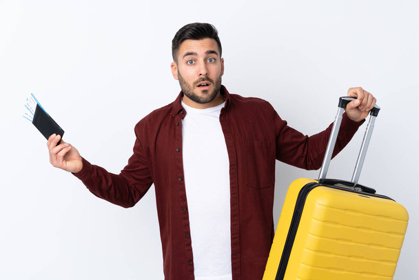 Young handsome man over isolated white background in vacation with suitcase and passport and surprised - Foto, afbeelding