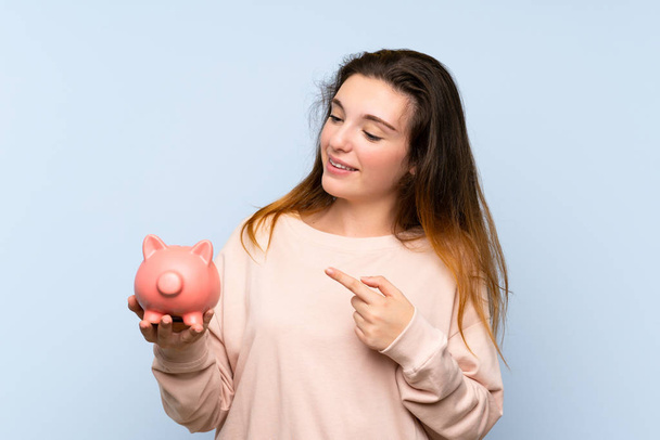 Young brunette girl over isolated blue background holding a big piggybank - 写真・画像
