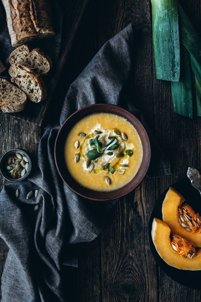 Гарбузовий крем-суп з вершками і гарбузовим насінням в красивій обстановці з гарбузовими шматочками, темним хлібом і цибулею-пореєм на темному тлі
 - Фото, зображення