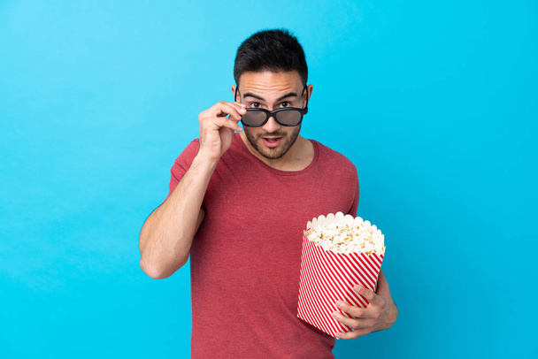 junger gutaussehender Mann über isoliertem blauem Hintergrund überrascht mit 3D-Brille und einem großen Eimer Popcorn - Foto, Bild