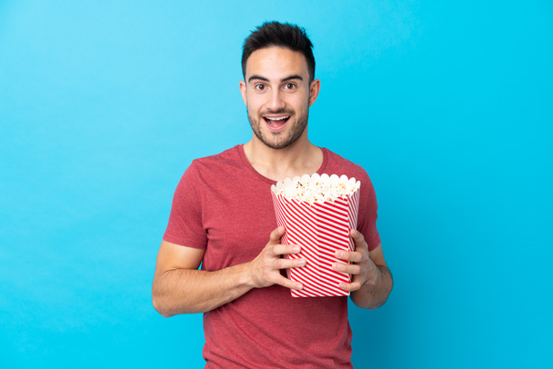 młody przystojny mężczyzna na odizolowanym niebieskim tle trzymając duży wiadro popcornów - Zdjęcie, obraz