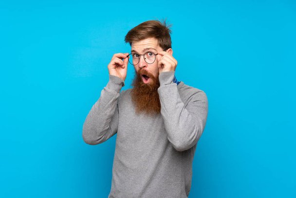 Zrzavý muž s dlouhým vousem přes izolované modré pozadí s brýlemi a překvapený - Fotografie, Obrázek