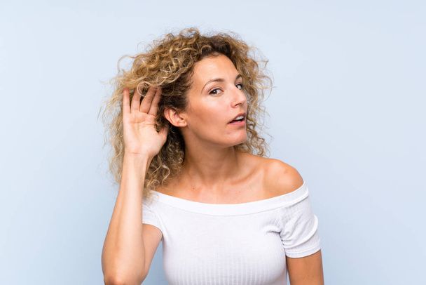 Young blonde woman with curly hair over isolated blue background listening something - Photo, Image