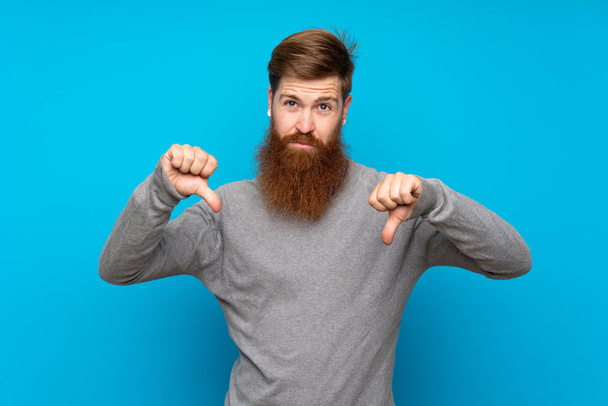 Redhead man with long beard over isolated blue background showing thumb down - 写真・画像