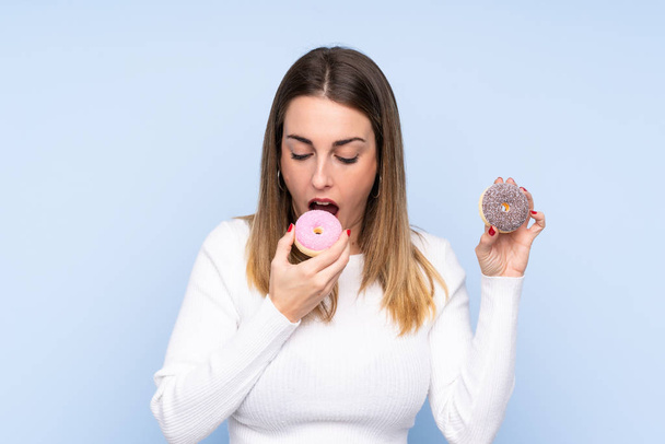 Young blonde woman over isolated blue background eating a donut - Фото, изображение