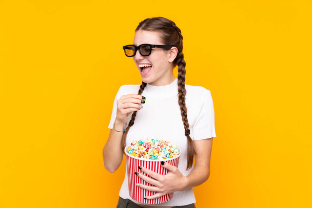 Young woman over isolated yellow background with 3d glasses and holding a big bucket of popcorns while pointing front - Zdjęcie, obraz