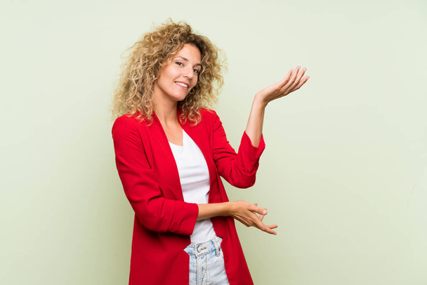 Young blonde woman with curly hair over isolated green background extending hands to the side for inviting to come - Фото, зображення