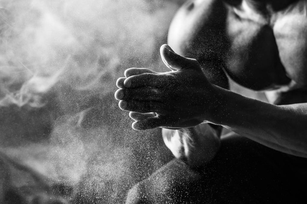 Bodybuilder uses hand magnesia. A man sits on a black background with smoke. Black and white shot - Фото, зображення