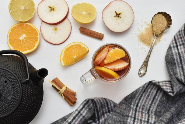 Θερμαινόμενο αρωματικό Χριστουγεννιάτικο τσάι με φρέσκα μήλα, πορτοκάλι, λεμόνι και κανέλα, επίπεδη θέσει με ζεστό κασκόλ και σίδερο κατσαρόλα τσάι. - Φωτογραφία, εικόνα